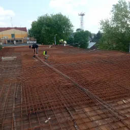 Prace zbrojarskie na budowie: robotnicy układają siatkę z prętów zbrojeniowych na deskowaniu, widok na budynek w tle i wieżę telekomunikacyjną.