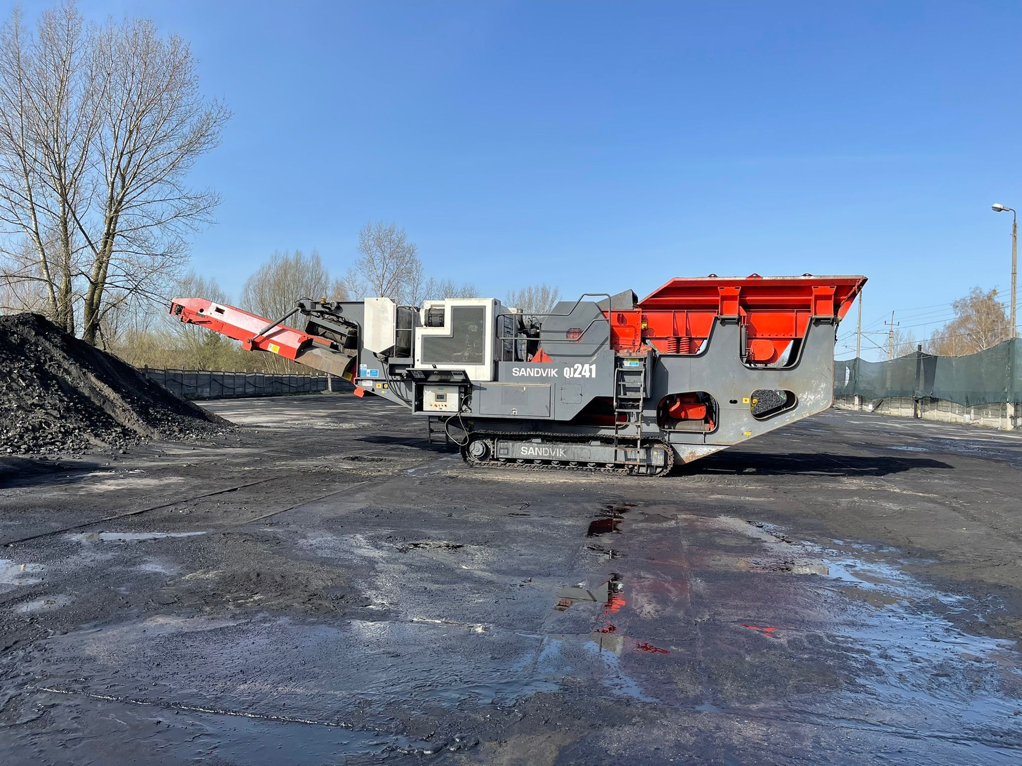 Mobilny przesiewacz Sandvik QJ241 na gąsienicach, z hałdą czarnego gruzu w tle, na placu pokrytym błotem i pyłem węglowym.