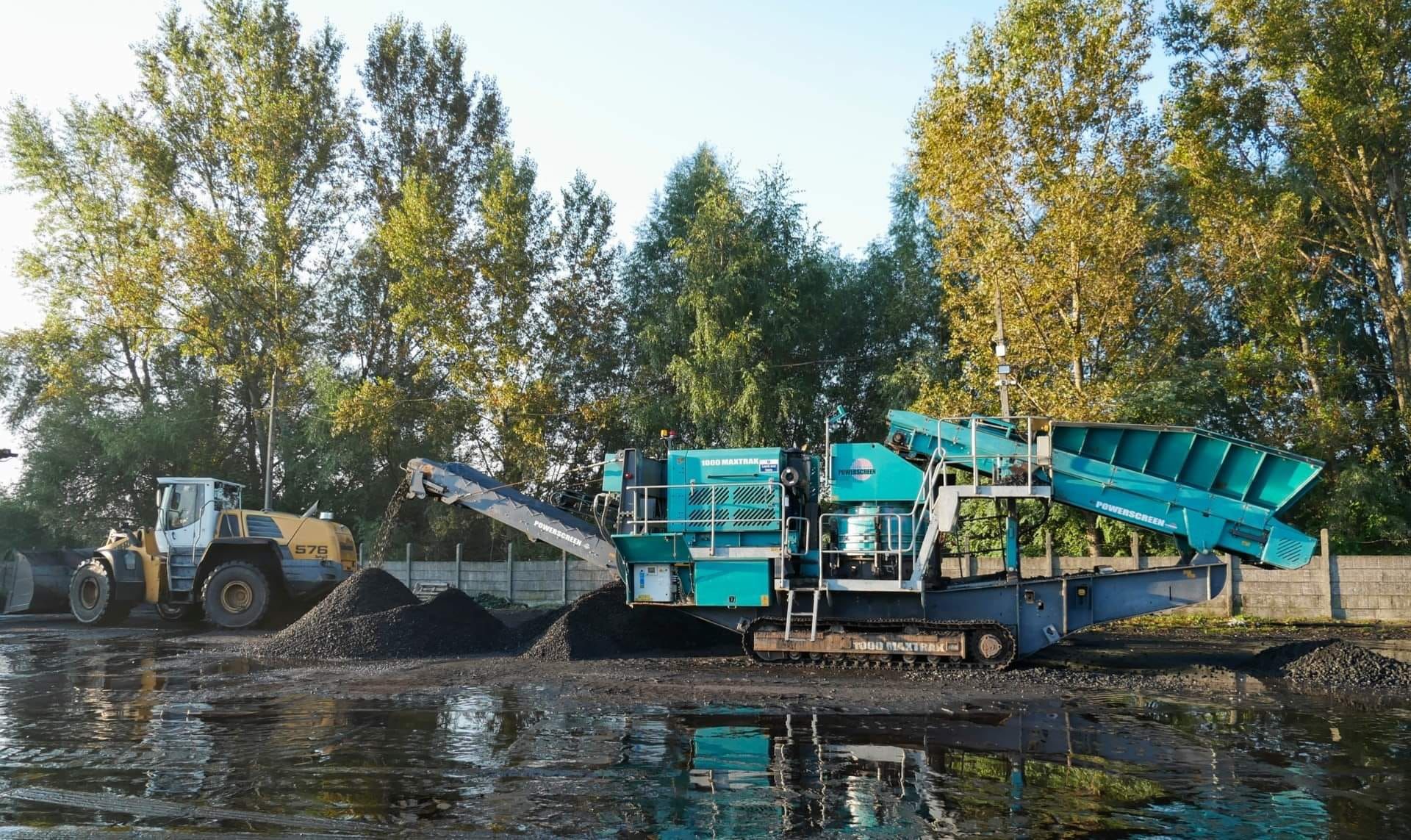 Turkusowa kruszarka Powerscreen 1000 Maxtrak na gąsienicach przerabia materiał na hałdę, obok żółta ładowarka Liebherr 576, teren podmokły, w tle drzewa.