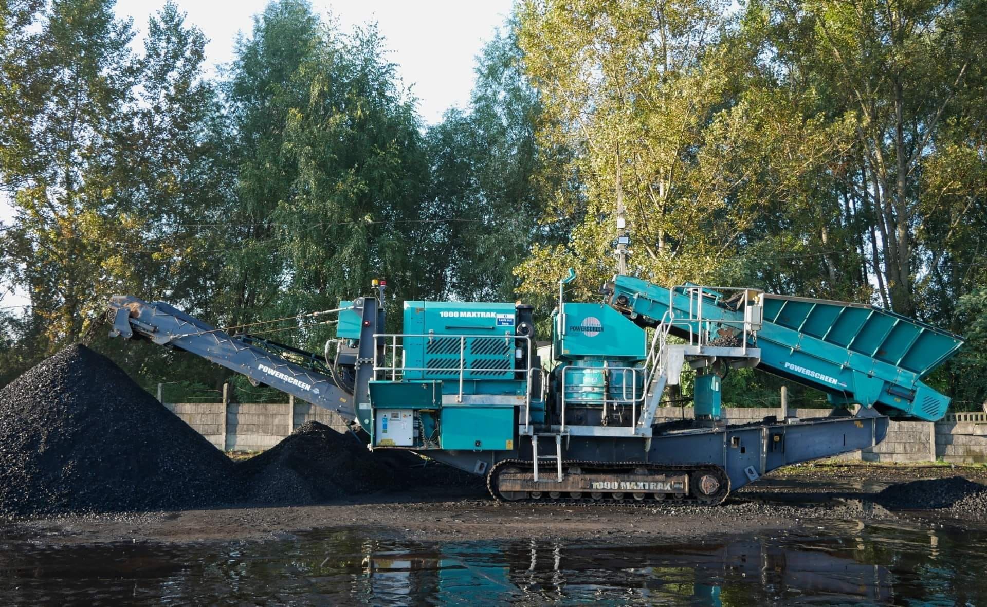Mobilny przesiewacz Powerscreen 1000 Maxtrak z podajnikiem, usypujący ciemny materiał na pryzmę w otoczeniu drzew i kałuży.