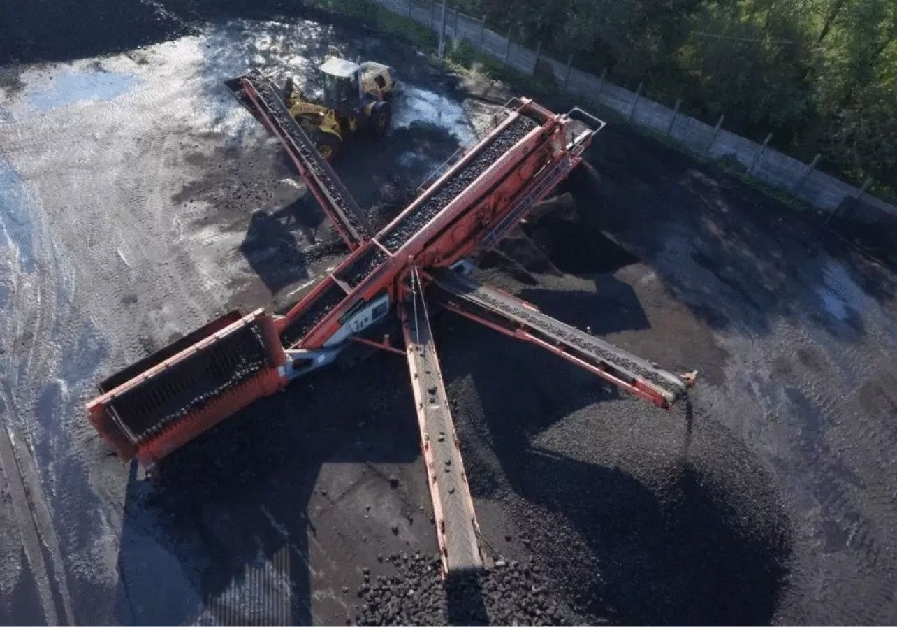 Widok z góry na rozbudowaną maszynę przesiewającą z kilkoma taśmociągami transportującymi ciemny materiał sypki, prawdopodobnie węgiel lub kamień, z żółtą ładowarką kołową w tle.
