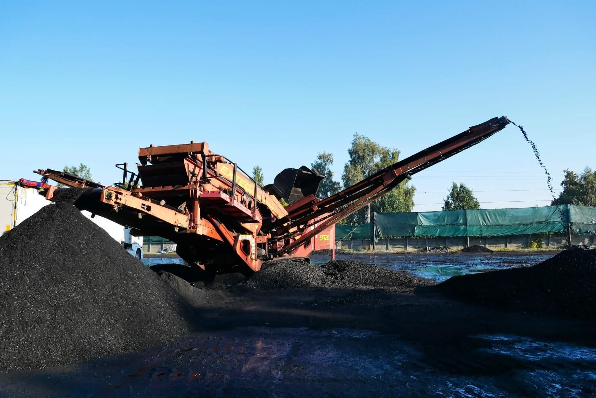 Pomarańczowa maszyna przesiewająca gruz z długim wysięgnikiem, formująca czarny kopiec na ziemi, widoczny fragment naczepy w tle.