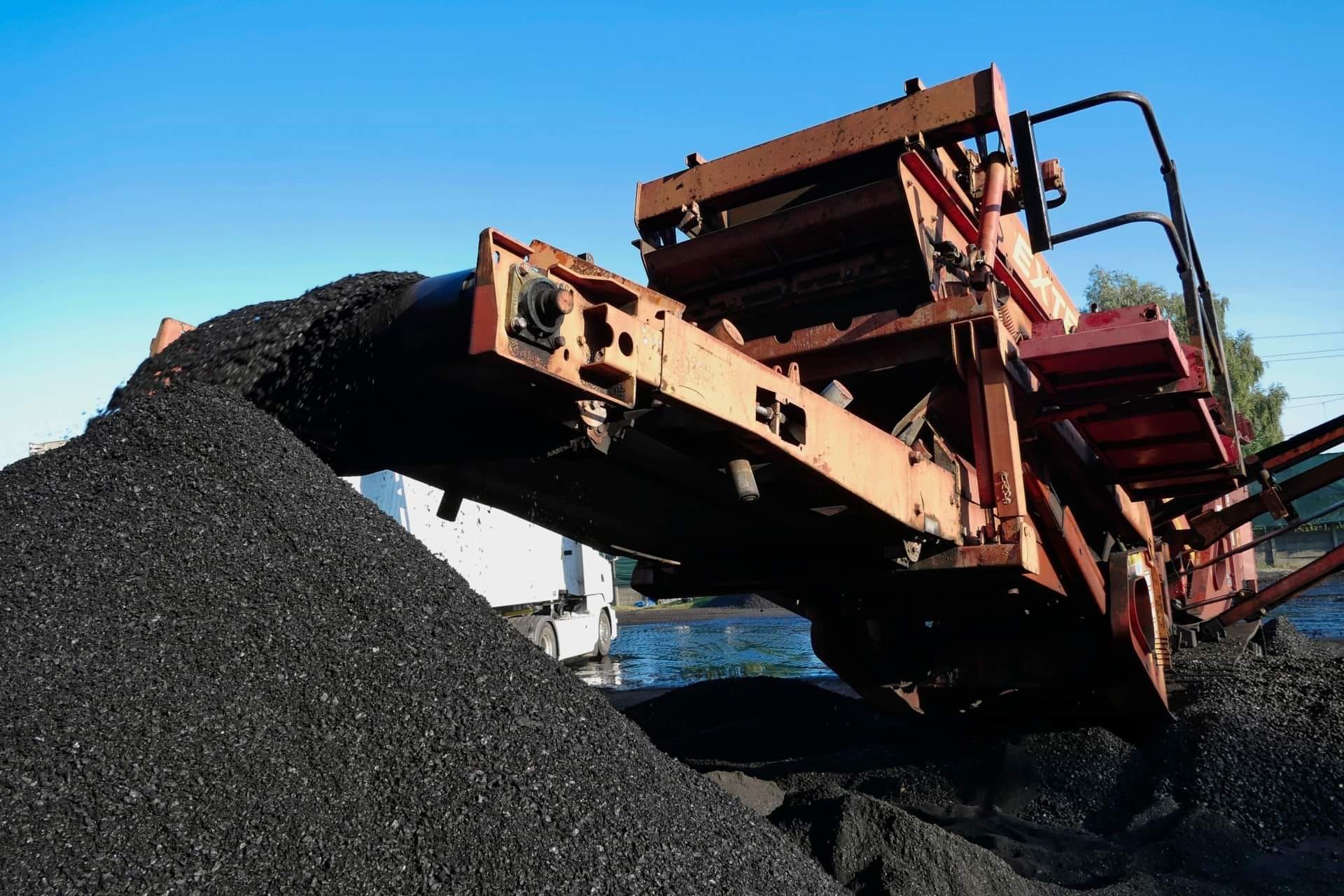 Pomarańczowa maszyna budowlana wysypuje czarny gruz na hałdę, widoczny fragment białej ciężarówki w tle.