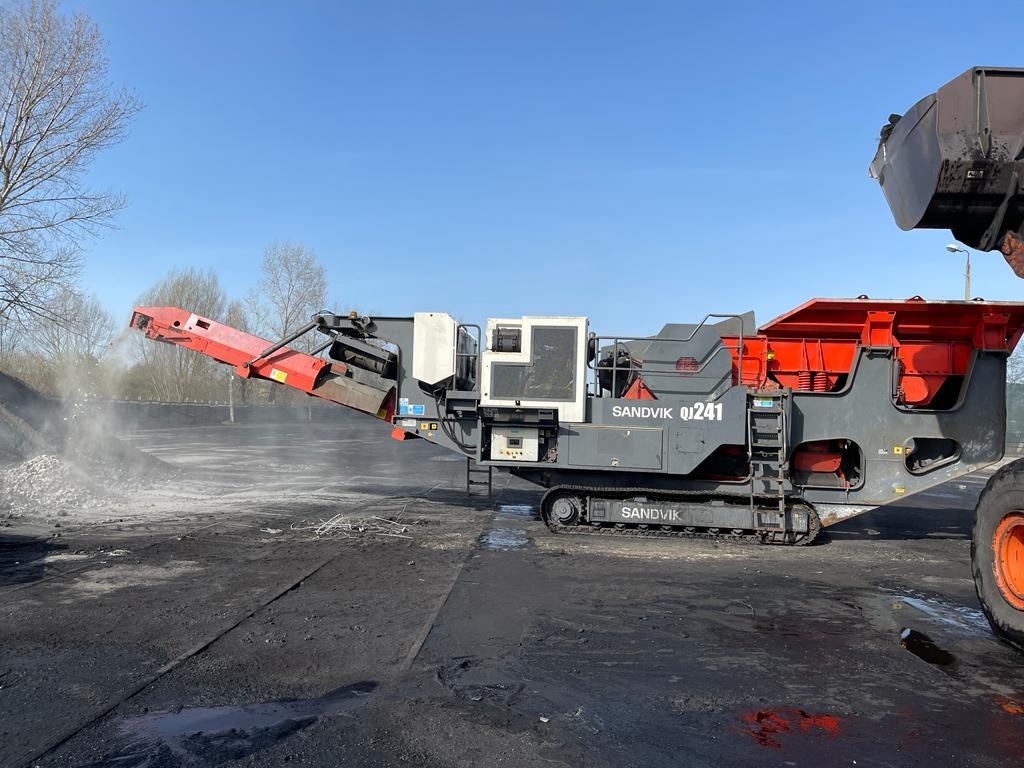 Mobilny przesiewacz kruszywa Sandvik QJ241 w trakcie pracy, z widocznym pyłem i odpadami, na tle szarego nieba i drzew bez liści.
