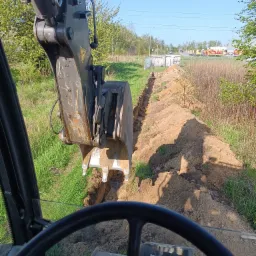 Widok z kabiny koparki na wykopany wąski rów w ziemi, z zielenią i budynkami w tle, wykonany w słoneczny dzień.