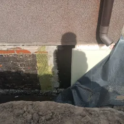 Odsłonięty fundament budynku z widoczną izolacją pionową, siatką zbrojeniową i folią ochronną, częściowo zasypany ziemią, z brązową rynną w rogu.