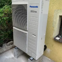 Zewnętrzna jednostka klimatyzacji Panasonic Inverter, umieszczona na betonowych podstawach przy żółtej ścianie budynku z widoczną roślinnością w tle.