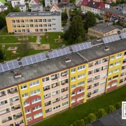 Widok z góry na blok mieszkalny z zainstalowanymi panelami słonecznymi na dachu, w tle budynek szkoły i plac zabaw, w prawym dolnym rogu logo firmy.