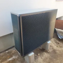 Zewnętrzna jednostka pompy ciepła Daikin, ustawiona na betonowych bloczkach przed białym budynkiem, widoczne przygotowania do instalacji w ziemi.