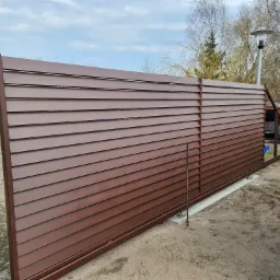 Brązowa, przesuwna brama lamelowa w trakcie montażu na betonowej podstawie, widoczna z boku, z elementami konstrukcyjnymi i osobą w tle.