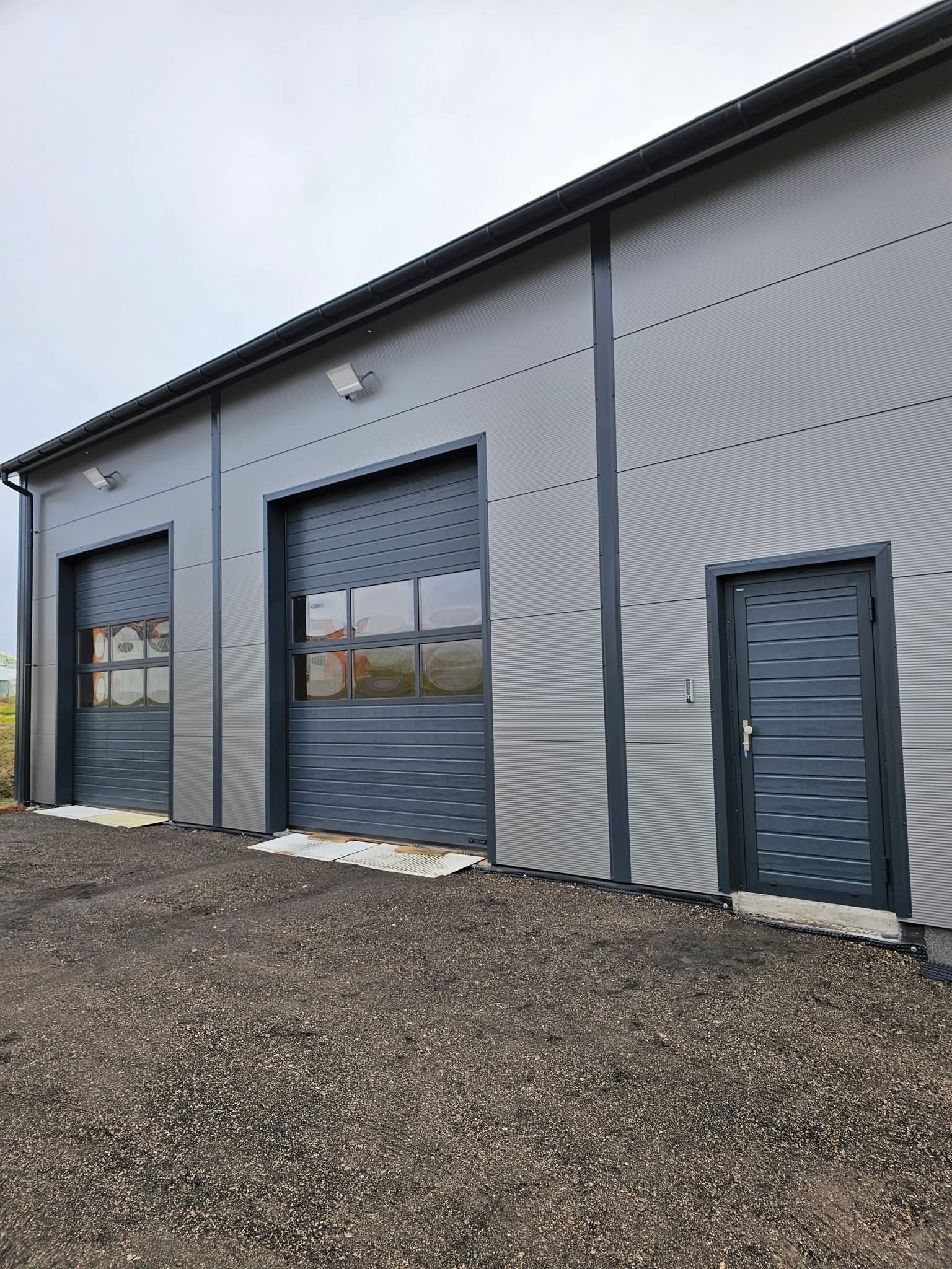 Szara hala z dwiema segmentowymi bramami garażowymi z oknami i drzwiami wejściowymi. Widok z zewnątrz na gotowy budynek z panelami.