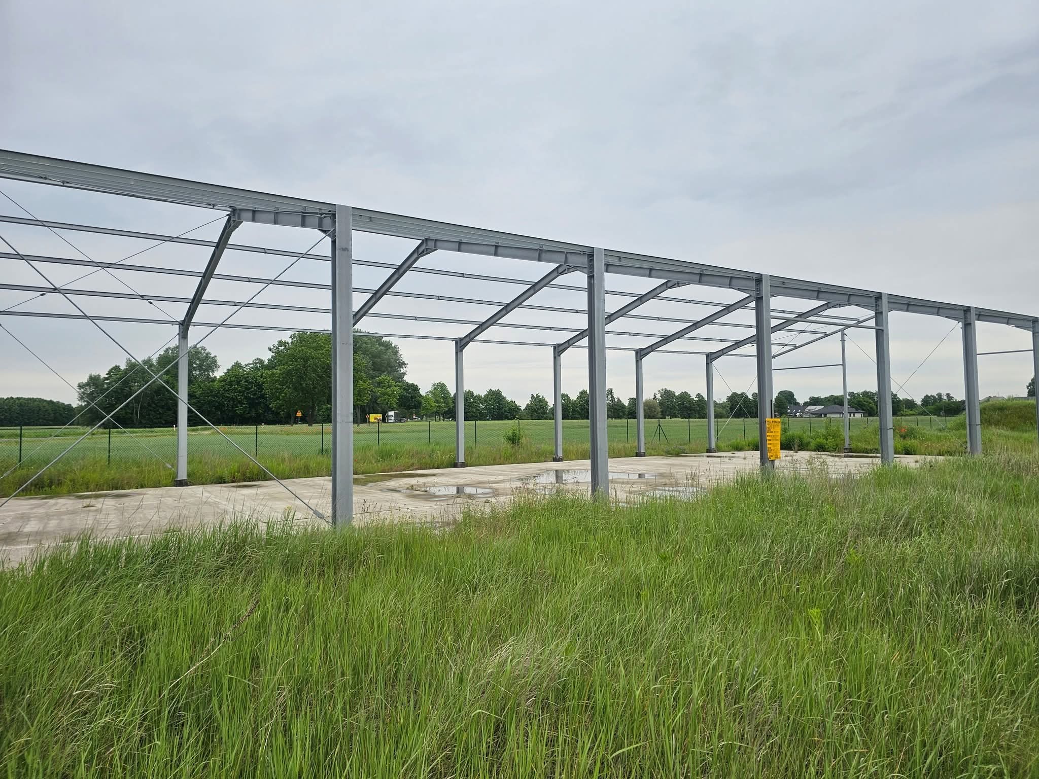 Stalowa konstrukcja hali w trakcie budowy, widoczna na tle zieleni i pochmurnego nieba. Elementy spawane, surowy beton fundamentu. Widok z poziomu gruntu.