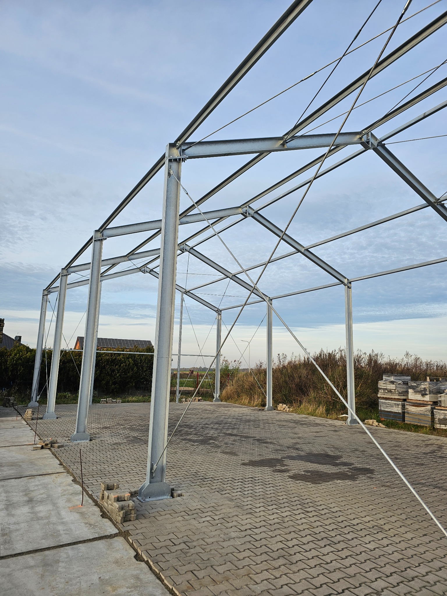 Stalowa konstrukcja hali w trakcie budowy, widok z dołu na szkielet z kolumn i belek, kostka brukowa na podłożu, niebo w tle. Widoczne detale łączeń i mocowań.