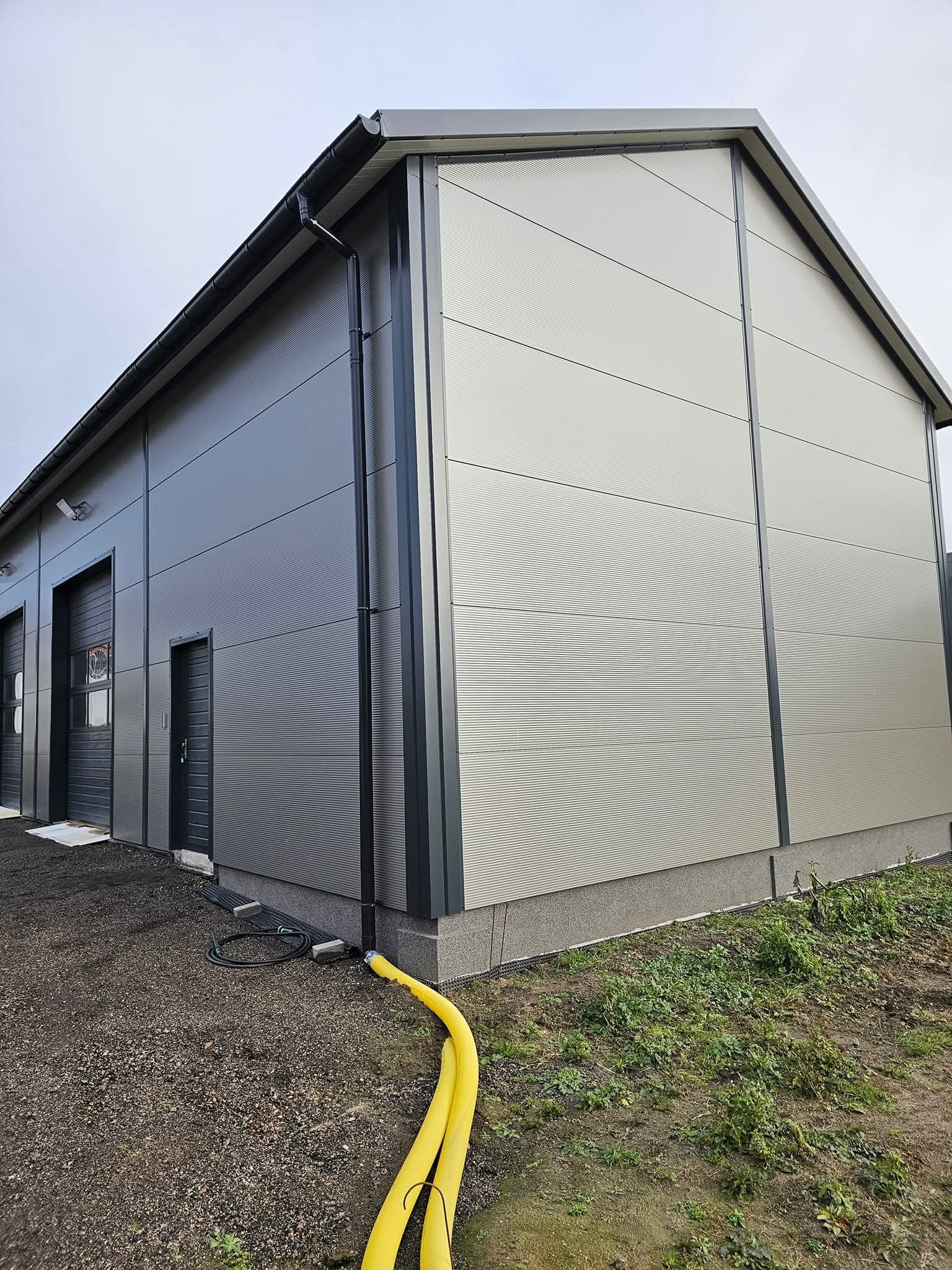 Nowoczesna hala z blachy trapezowej w odcieniach szarości, z widocznymi drzwiami garażowymi i wejściowymi. Uwagę zwraca żółty wąż na ziemi.