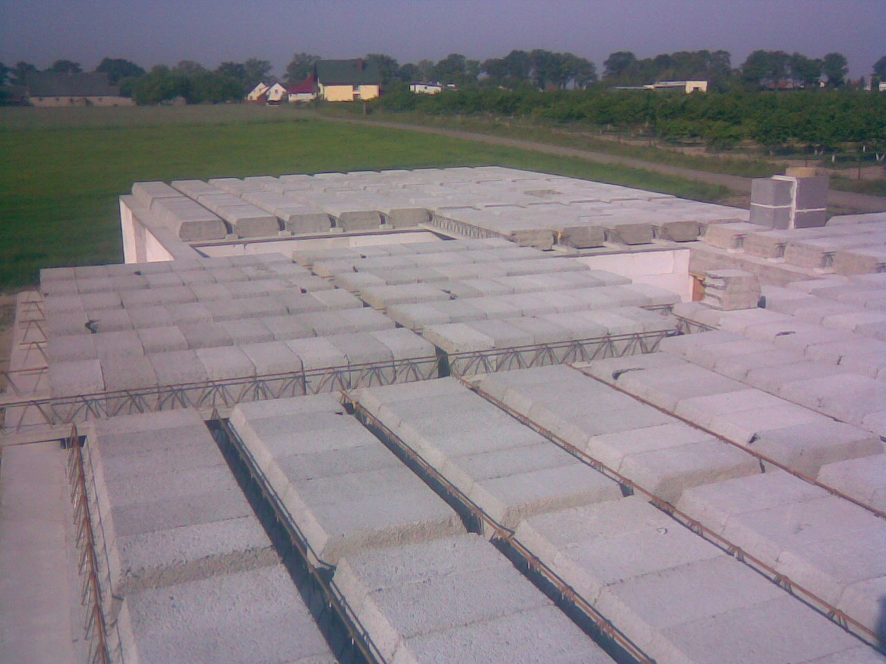 Wznoszenie stropu Teriva z belek kratownicowych i betonowych pustaków na budowie domu jednorodzinnego na tle zielonego pola i zabudowań wiejskich.