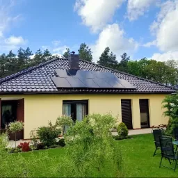 Dom jednorodzinny z brązową dachówką i panelami słonecznymi na dachu, widok z ogrodu z zielonym trawnikiem i ozdobnymi krzewami.