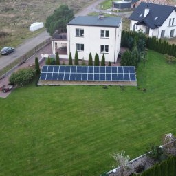 ESOLEO Sp. z o.o. - Widok z góry na panele słoneczne zainstalowane na trawniku przed domem jednorodzinnym, z widocznym samochodem na drodze i przyczepką.