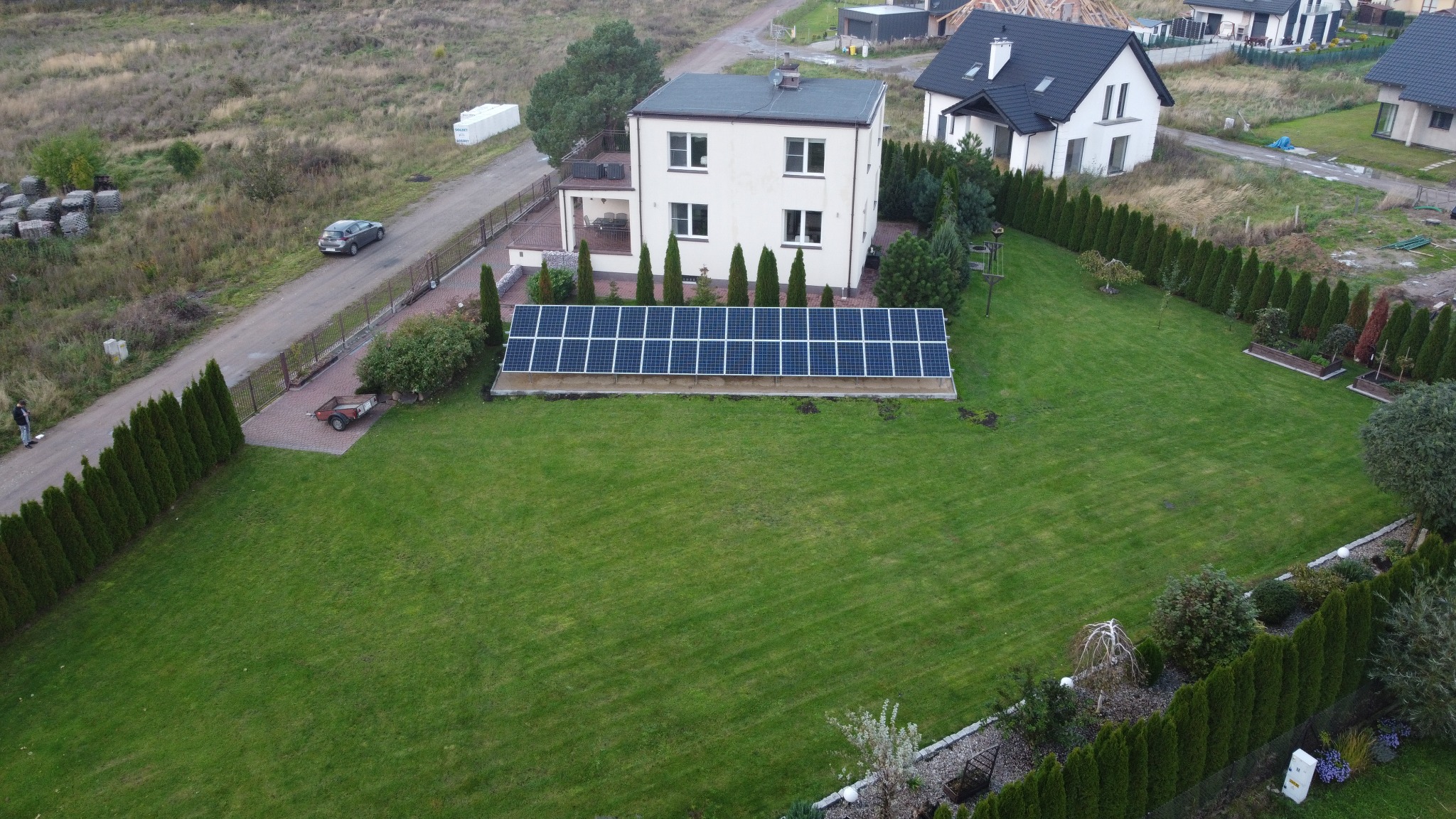 Widok z góry na panele słoneczne zainstalowane na trawniku przed domem jednorodzinnym, z widocznym samochodem na drodze i przyczepką.