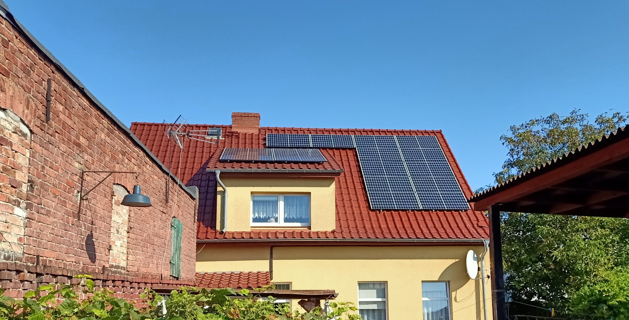 Panele słoneczne na dachu domu z czerwoną dachówką, widoczne również antena, komin i fragment ceglanego budynku obok.