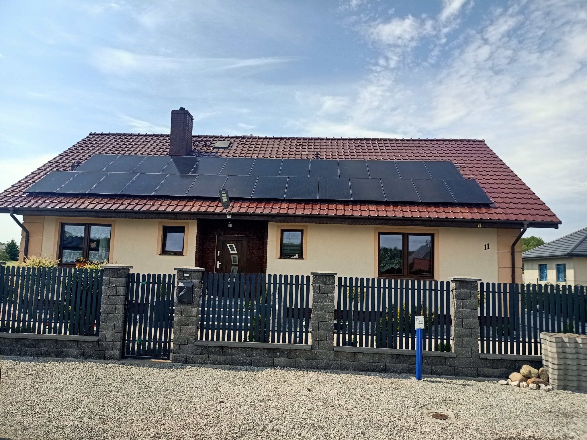 Dom jednorodzinny z brązowym dachem i panelami słonecznymi na połaci dachu, widok z poziomu gruntu, szary płot z betonowymi słupkami.