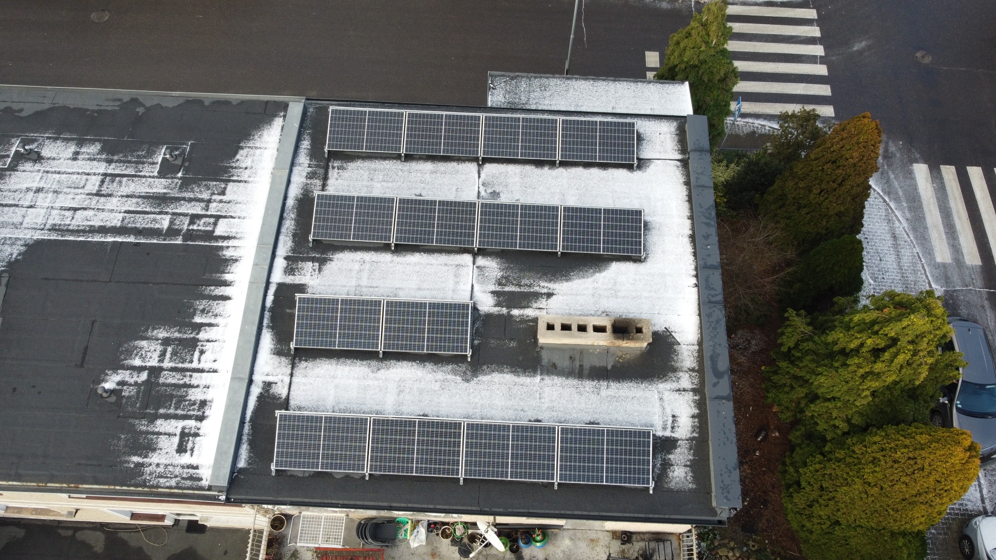 Widok z góry na częściowo ośnieżony dach budynku z zainstalowanymi panelami słonecznymi; w tle przejście dla pieszych i zieleń.