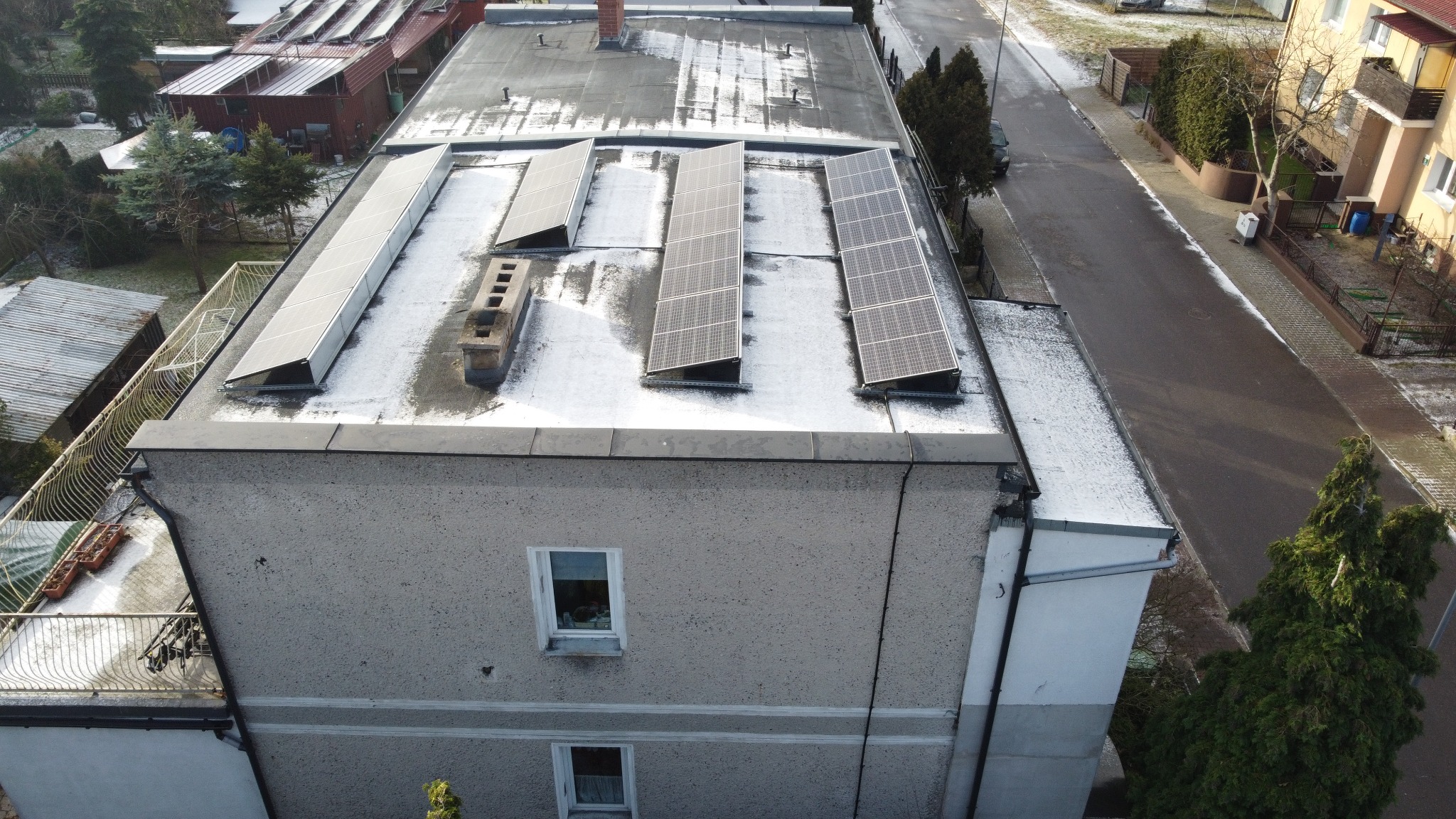 Panele słoneczne na płaskim dachu budynku mieszkalnego, częściowo pokryte śniegiem, widok z góry na ulicę i sąsiednie budynki.