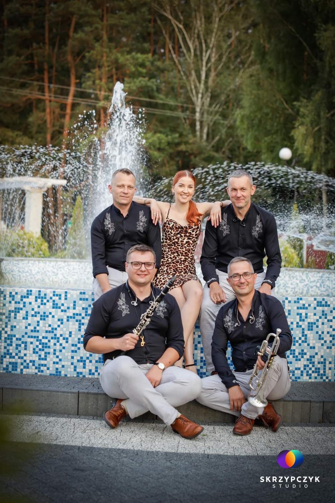Pięcioosobowa grupa wokalno-instrumentalna pozuje na tle fontanny: trzech mężczyzn w czarnych koszulach z haftem, jeden z klarnetem, drugi z trąbką, oraz kobieta w sukience w panterkę.