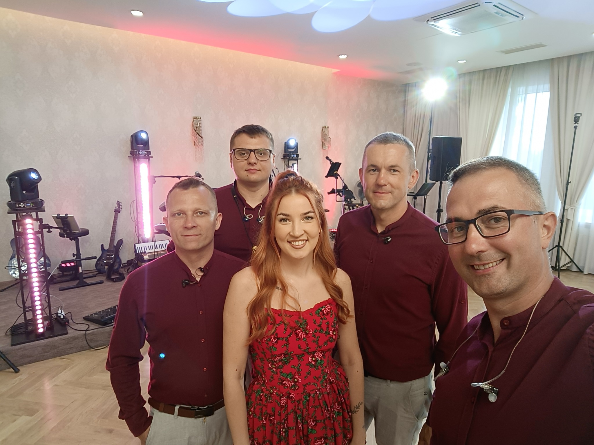 Zespół muzyczny w bordowych koszulach i wokalistka w czerwonej sukience w kwiaty, ustawieni na tle sceny z oświetleniem i instrumentami, ujęcie selfie.