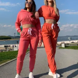 Dwie kobiety w dopasowanych dresach (różowy i pomarańczowy), stojące na tle jeziora i błękitnego nieba, prezentujące sportowy styl odzieży.