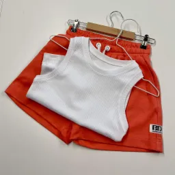 Zestaw letni: pomarańczowe szorty z logo i biała prążkowana koszulka na wieszaku ze spinaczami.