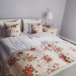 Łóżko z białą pościelą i narzutą w kwiaty, ozdobne poduszki, lampka nocna na szafce przy ścianie w odcieniach szarości.