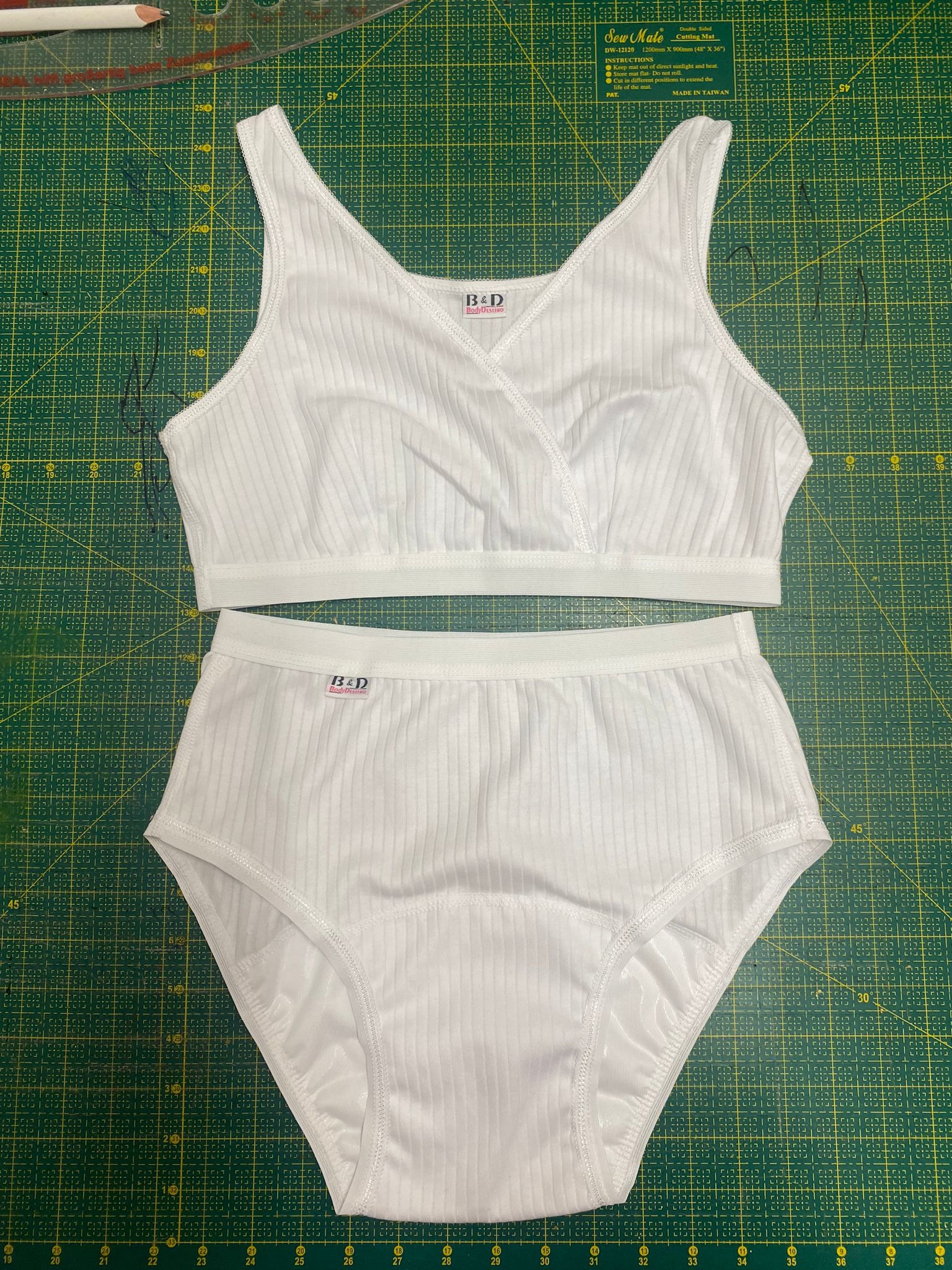 Biały, prążkowany komplet bielizny damskiej (stanik i majtki) z metką B&D, leżący na zielonej macie do cięcia z podziałką centymetrową, obok ołówek i linijka.