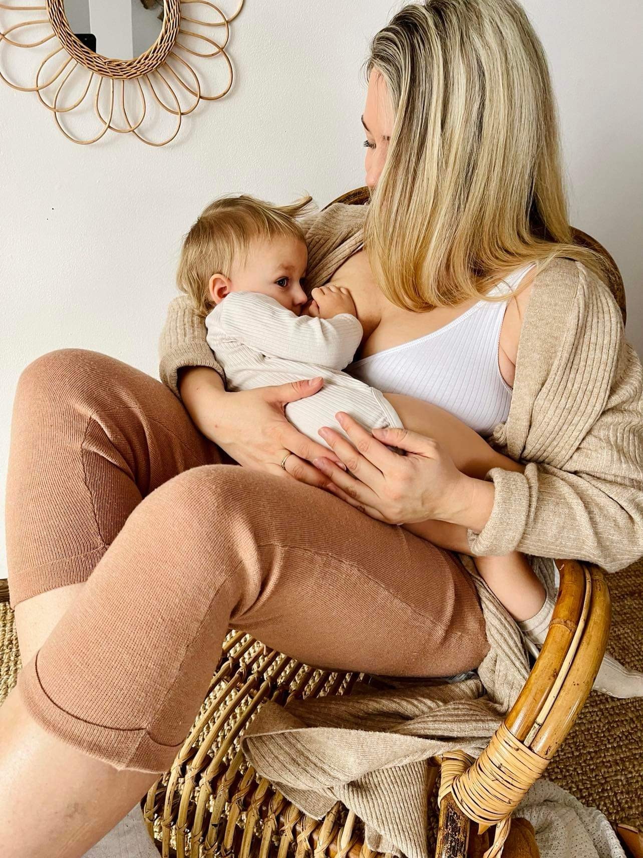 Kobieta karmiąca piersią dziecko ubranego w beżowe body na wiklinowym fotelu, w tle okrągłe lustro z wikliny.
