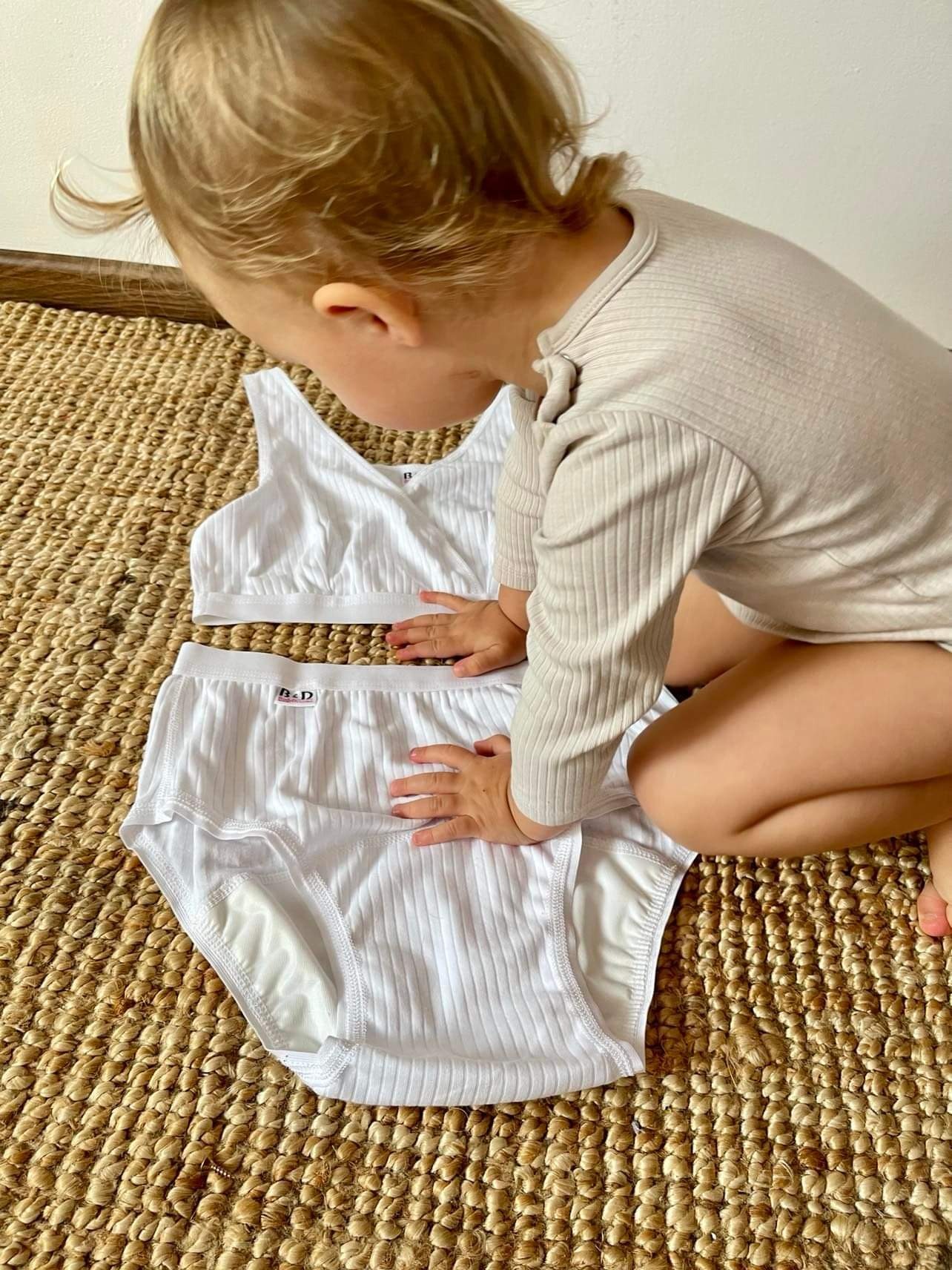 Dziecko dotyka dłońmi białego, prążkowanego kompletu bielizny (majtki i top) z metką B&D, leżącego na brązowym, plecionym dywaniku.