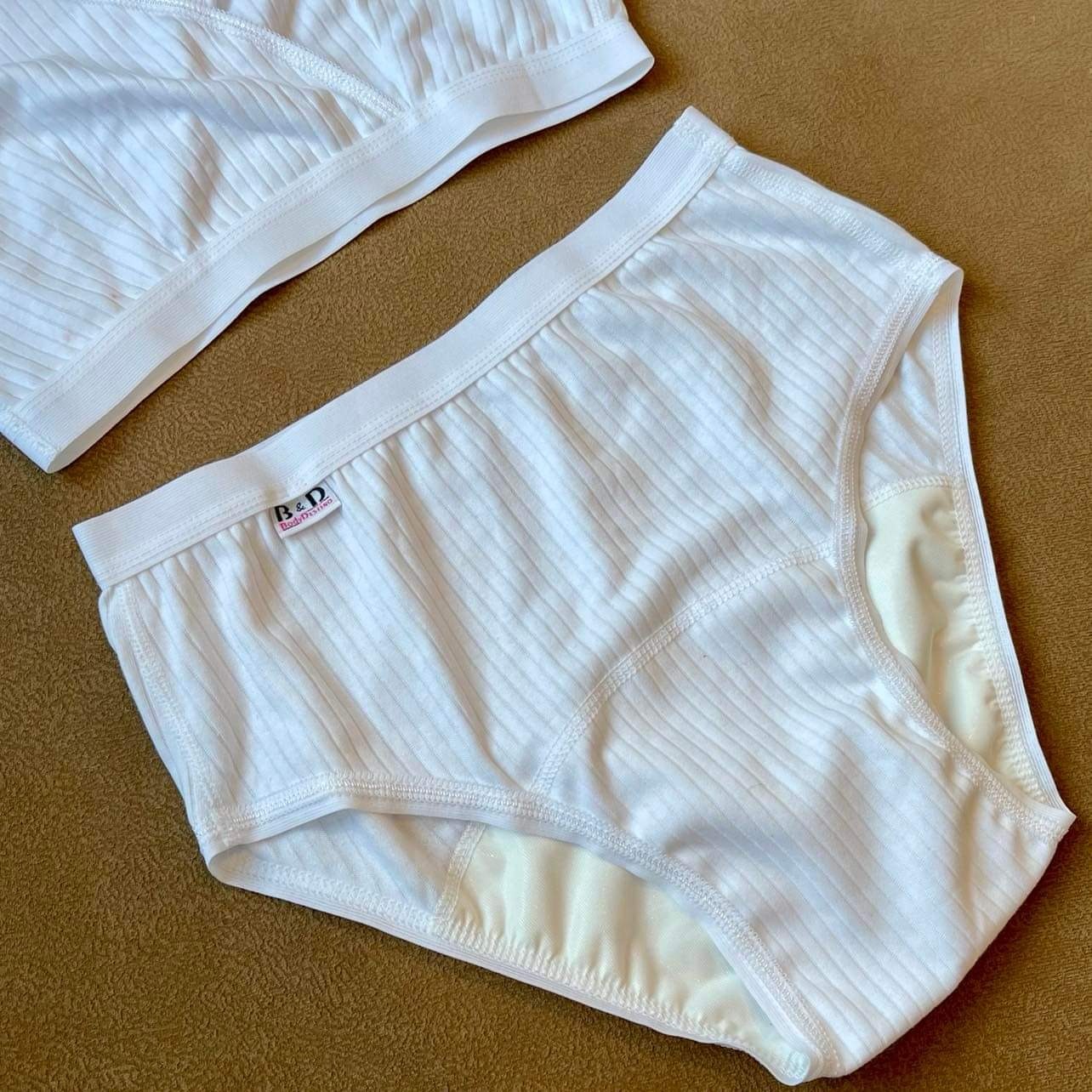 Biała, prążkowana bielizna damska z logo 'B&D BodyDressing' na brązowym tle.