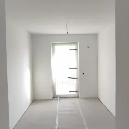 Pusty pokój w trakcie remontu, świeżo pomalowane białe ściany, zabezpieczona podłoga, widoczne ślady po pracach wykończeniowych.