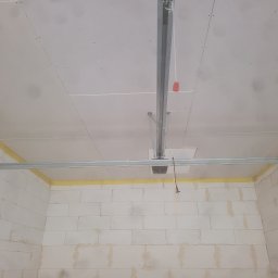Usługi Remontowo budowlane Mateusz Kwiecień - Wnętrze garażu w trakcie wykańczania: surowa ściana z białych bloczków, częściowo zakryta żółtą taśmą malarską, sufit z płyt kartonowo-gipsowych z widoczną konstrukcją metalową i mechanizmem bramy...