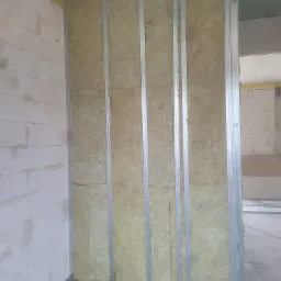 Metalowy szkielet ściany działowej wypełniony wełną mineralną, widoczny fragment ściany z bloczków betonowych i żółta folia izolacyjna przy podłodze.