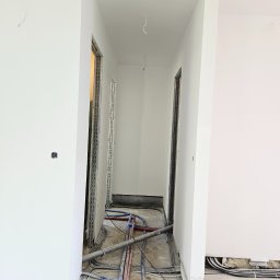 Usługi Remontowo budowlane Mateusz Kwiecień - Korytarz w trakcie remontu, widoczne przewody elektryczne i rury instalacyjne na podłodze, ściany pomalowane na biało, ościeżnice drzwiowe w trakcie montażu.