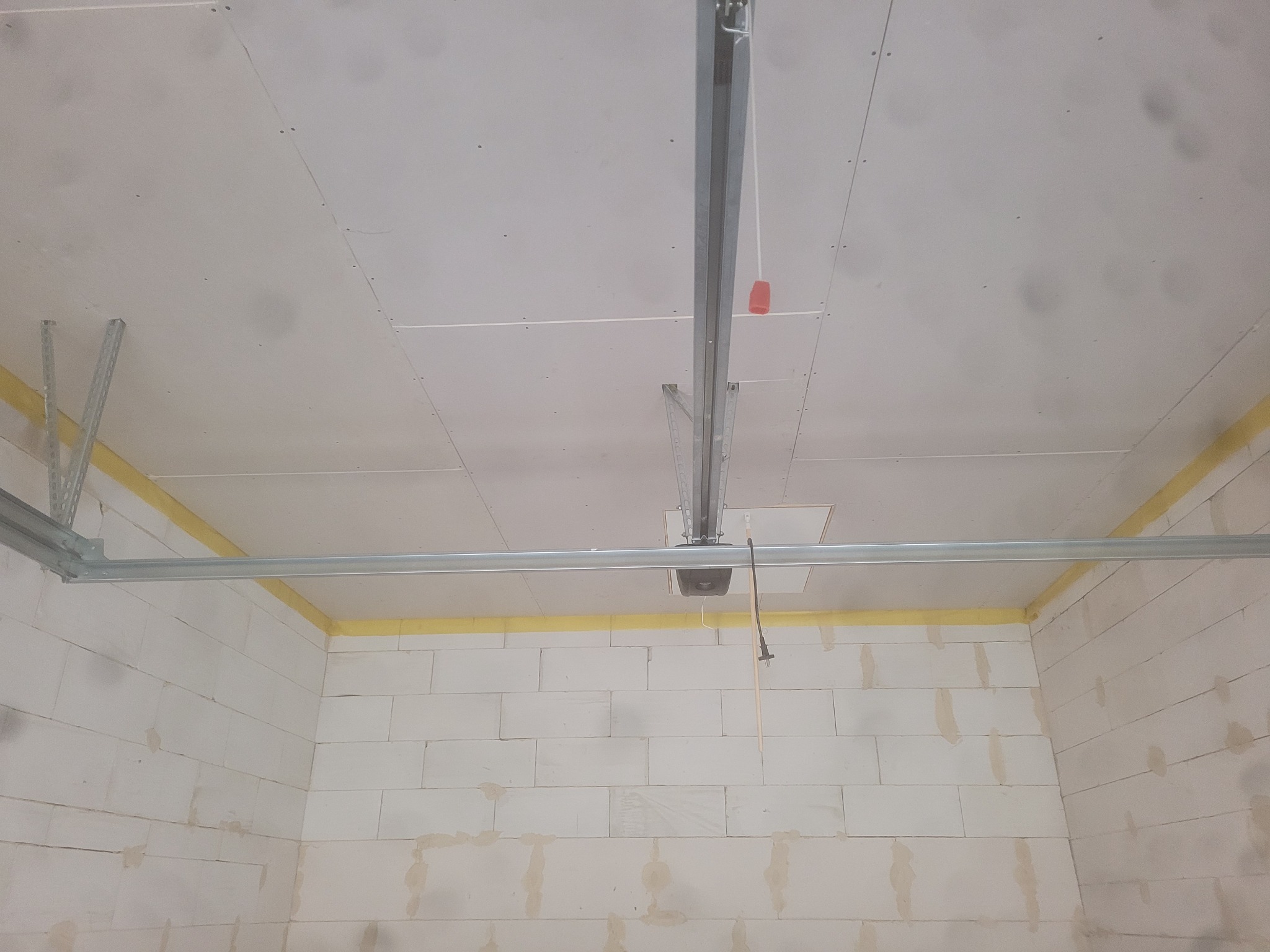 Wnętrze garażu w trakcie wykańczania: surowa ściana z białych bloczków, częściowo zakryta żółtą taśmą malarską, sufit z płyt kartonowo-gipsowych z widoczną konstrukcją metalową i mechanizmem bramy...