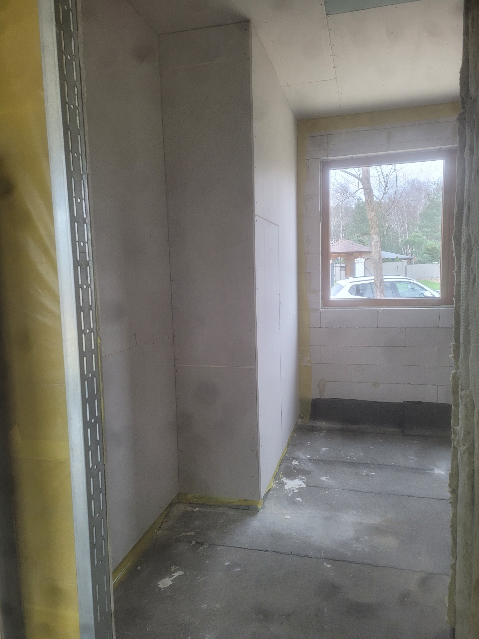 Wnętrze w trakcie remontu: ściany wyłożone płytami kartonowo-gipsowymi, widoczne profile metalowe, okno z widokiem na zewnątrz, surowy beton na podłodze.