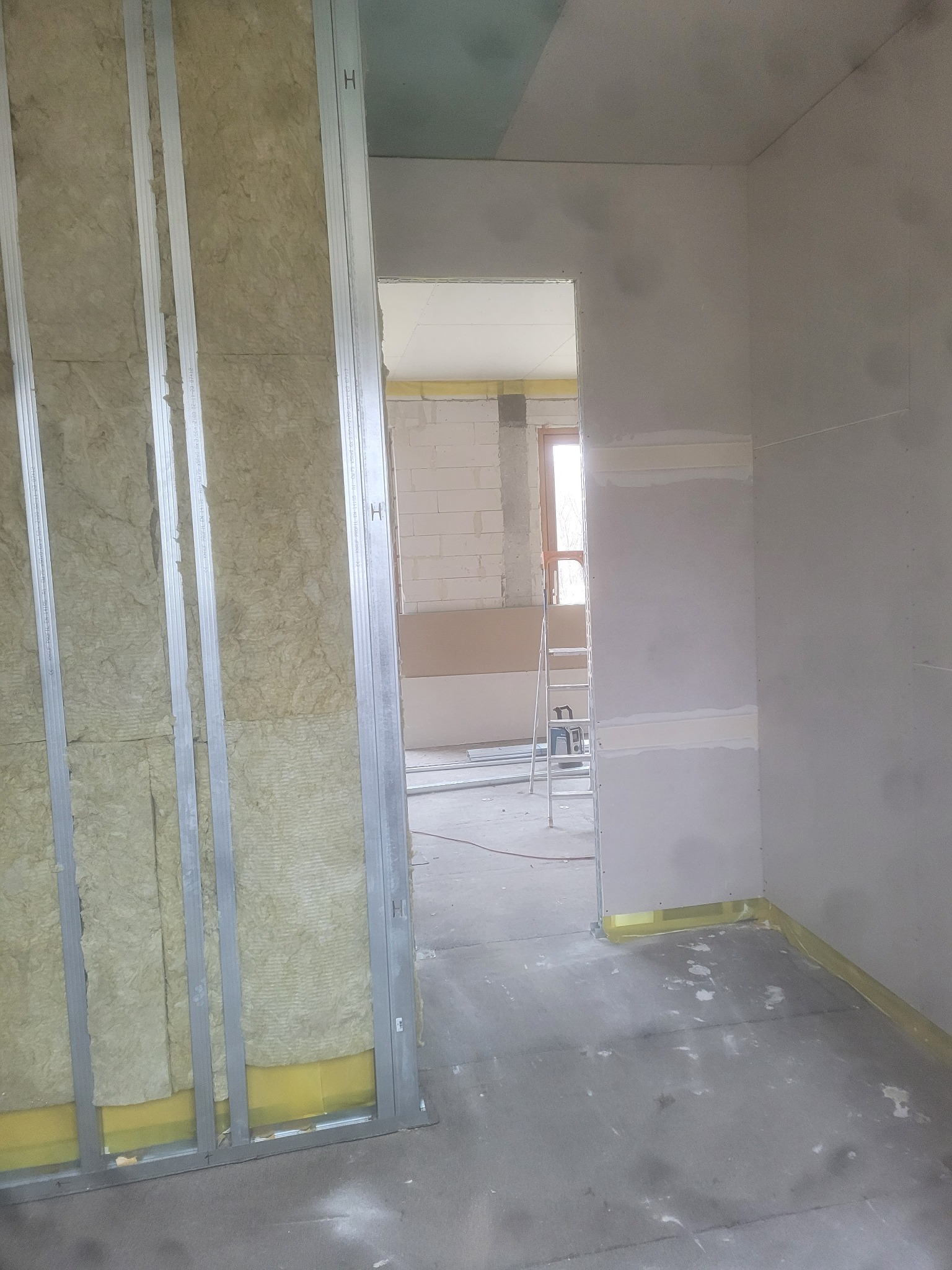 Wnętrze w trakcie prac wykończeniowych: metalowy stelaż z wełną mineralną, fragment ściany z płyt kartonowo-gipsowych z widocznymi łączeniami, surowy betonowy podkład, drabina w tle.