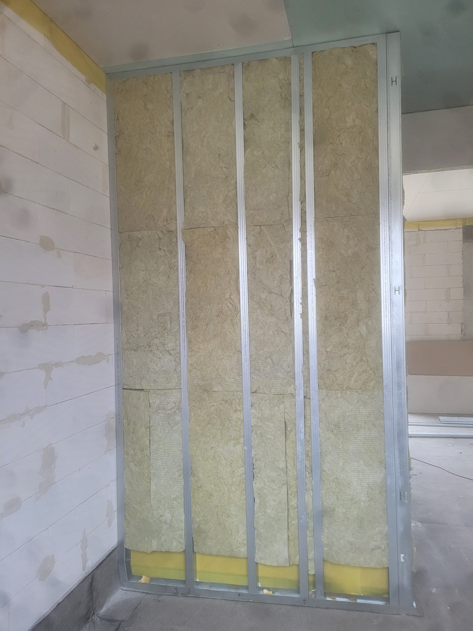 Metalowy szkielet ściany działowej wypełniony wełną mineralną, widoczny fragment ściany z bloczków betonowych i żółta folia izolacyjna przy podłodze.