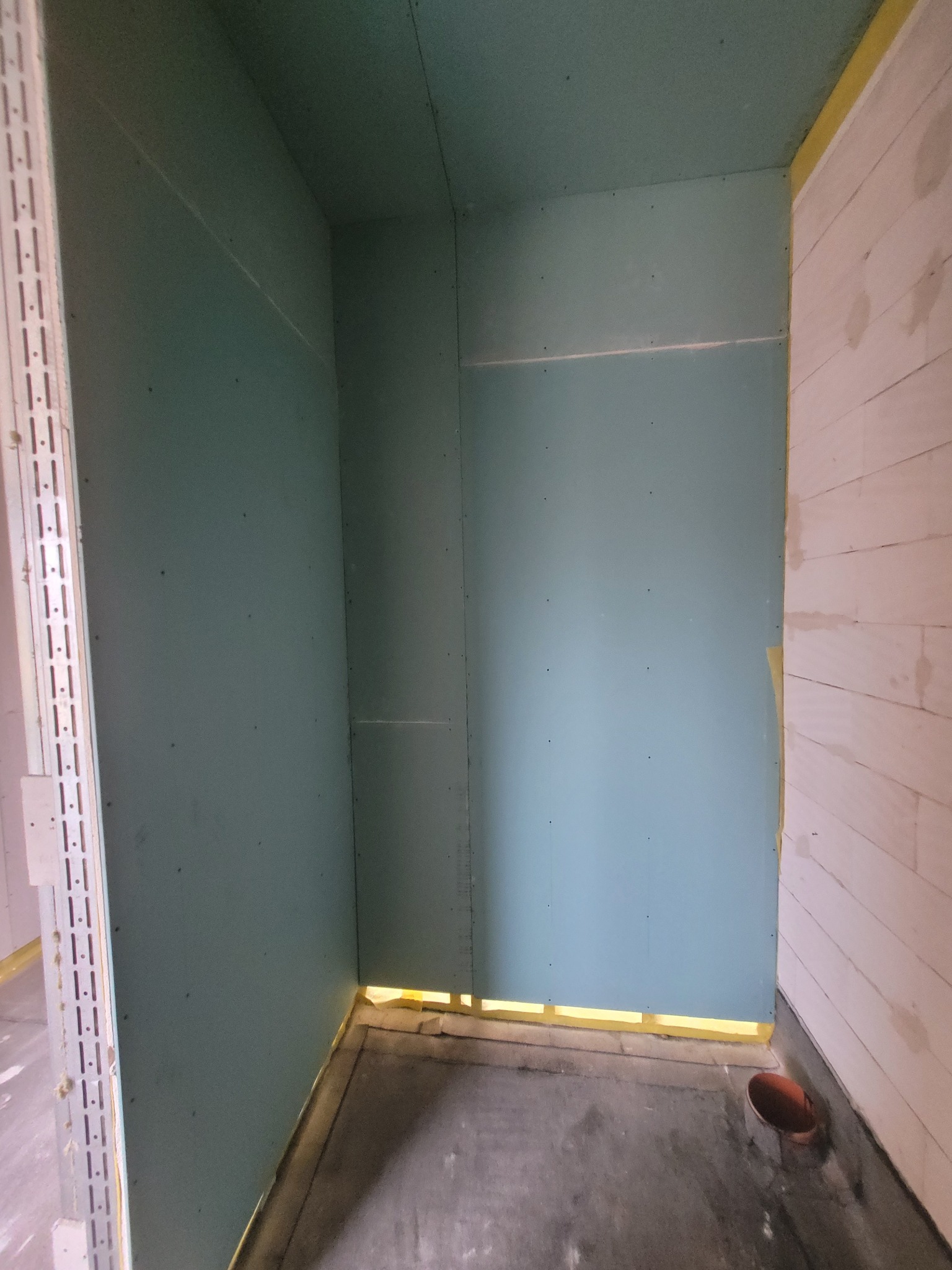 Wnętrze w trakcie prac wykończeniowych: ściany obłożone zielonymi płytami gipsowo-kartonowymi, widoczne łączenia i szpachlowania, surowy beton na podłodze, żółta taśma zabezpieczająca przy podłodze...