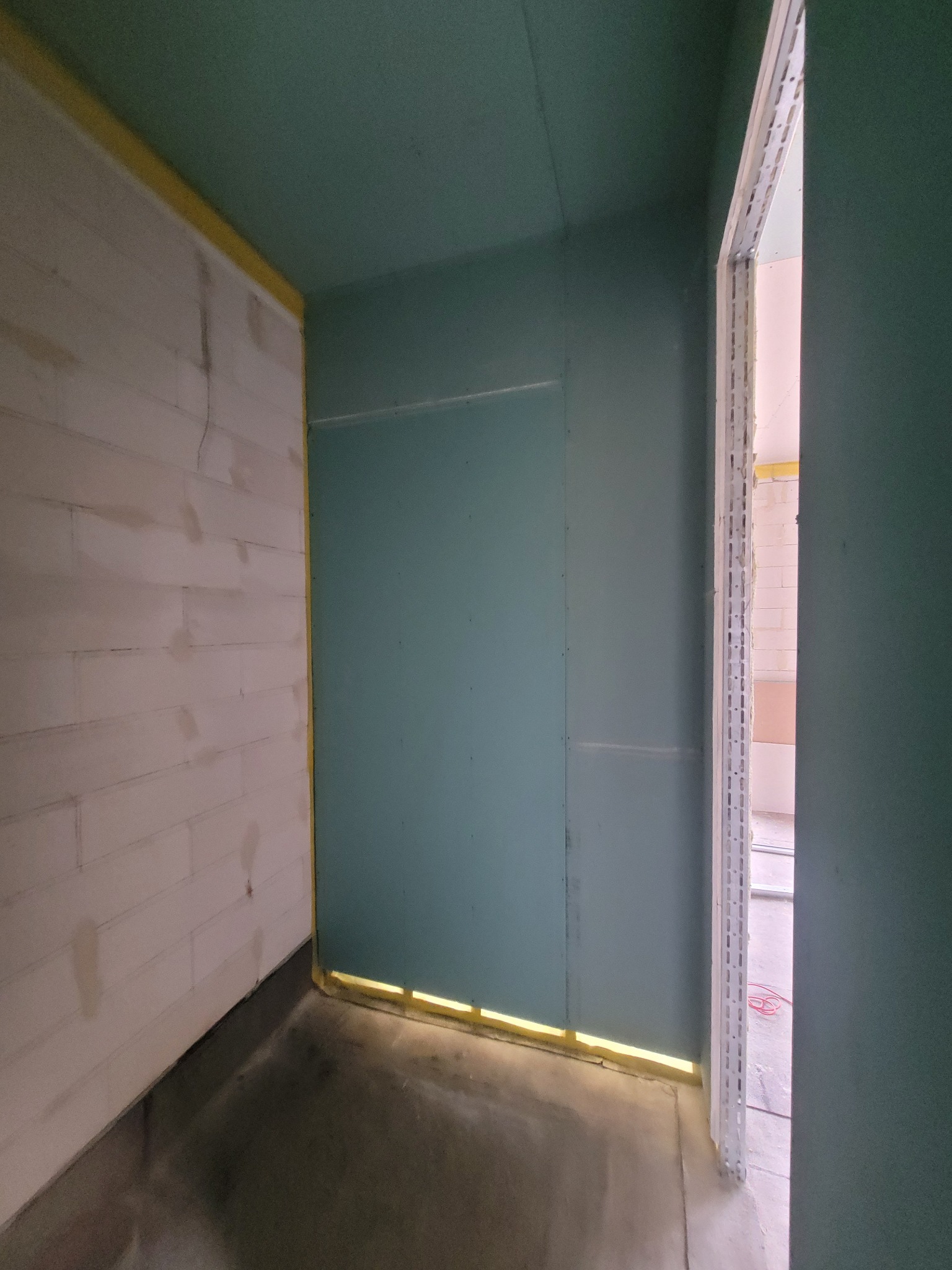 Wnętrze w trakcie prac wykończeniowych: ściany z płyt kartonowo-gipsowych, surowy beton na podłodze, widoczne profile ościeżnic drzwiowych, żółta taśma maskująca wzdłuż krawędzi.