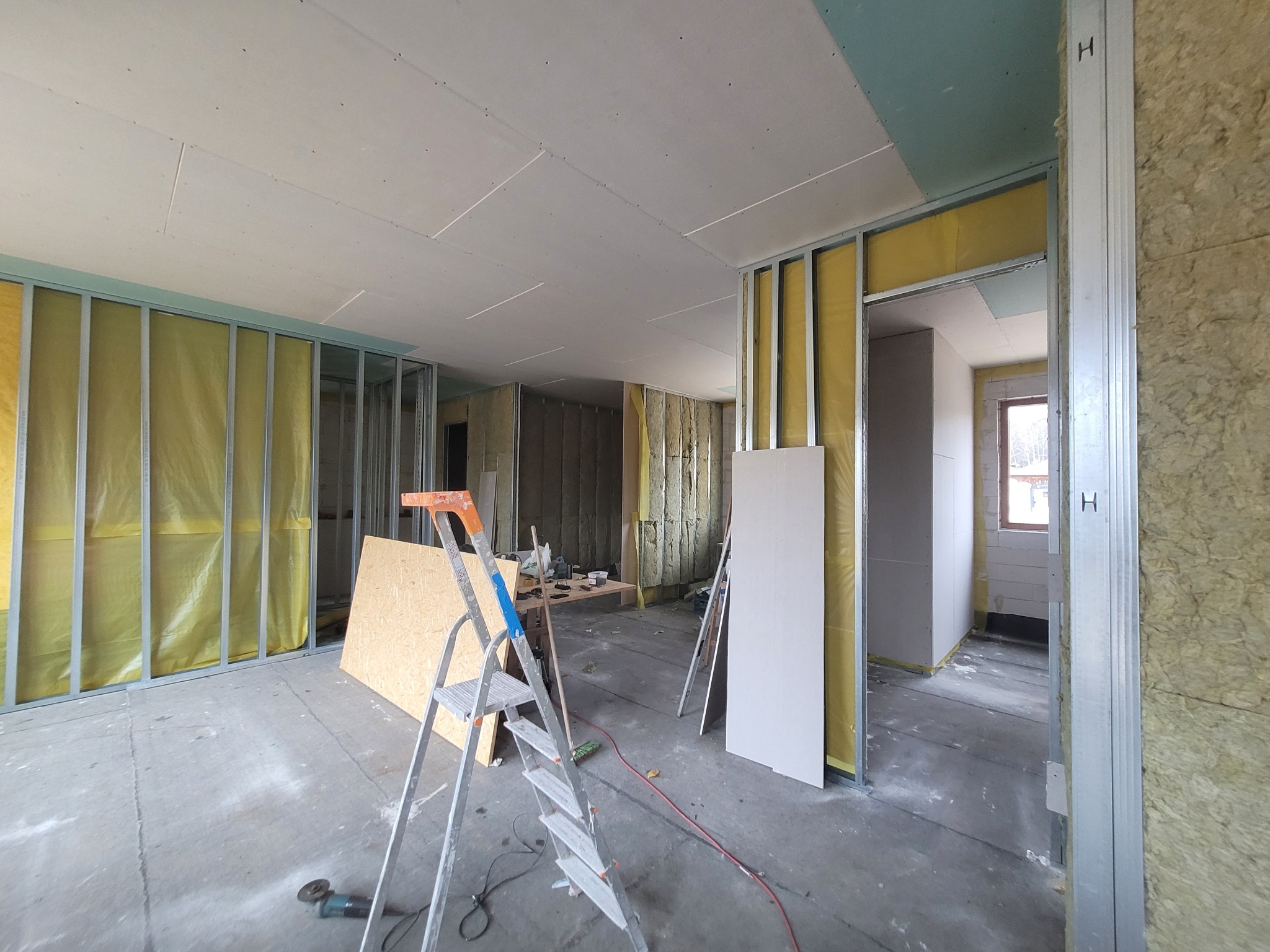 Wnętrze w trakcie budowy ścian działowych z metalowych profili wypełnionych wełną mineralną, częściowo obłożonych płytami gipsowo-kartonowymi; widoczna drabina, narzędzia i płyta OSB.
