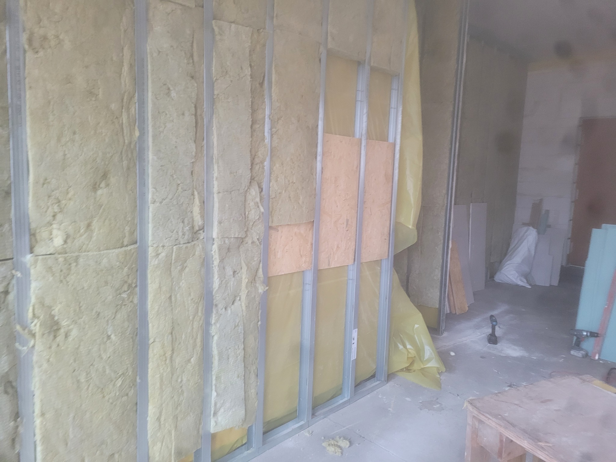 Montaż izolacji z wełny mineralnej w metalowym stelażu ściany działowej, częściowo zakrytej folią ochronną i płytami drewnianymi, widoczne narzędzia na podłodze.