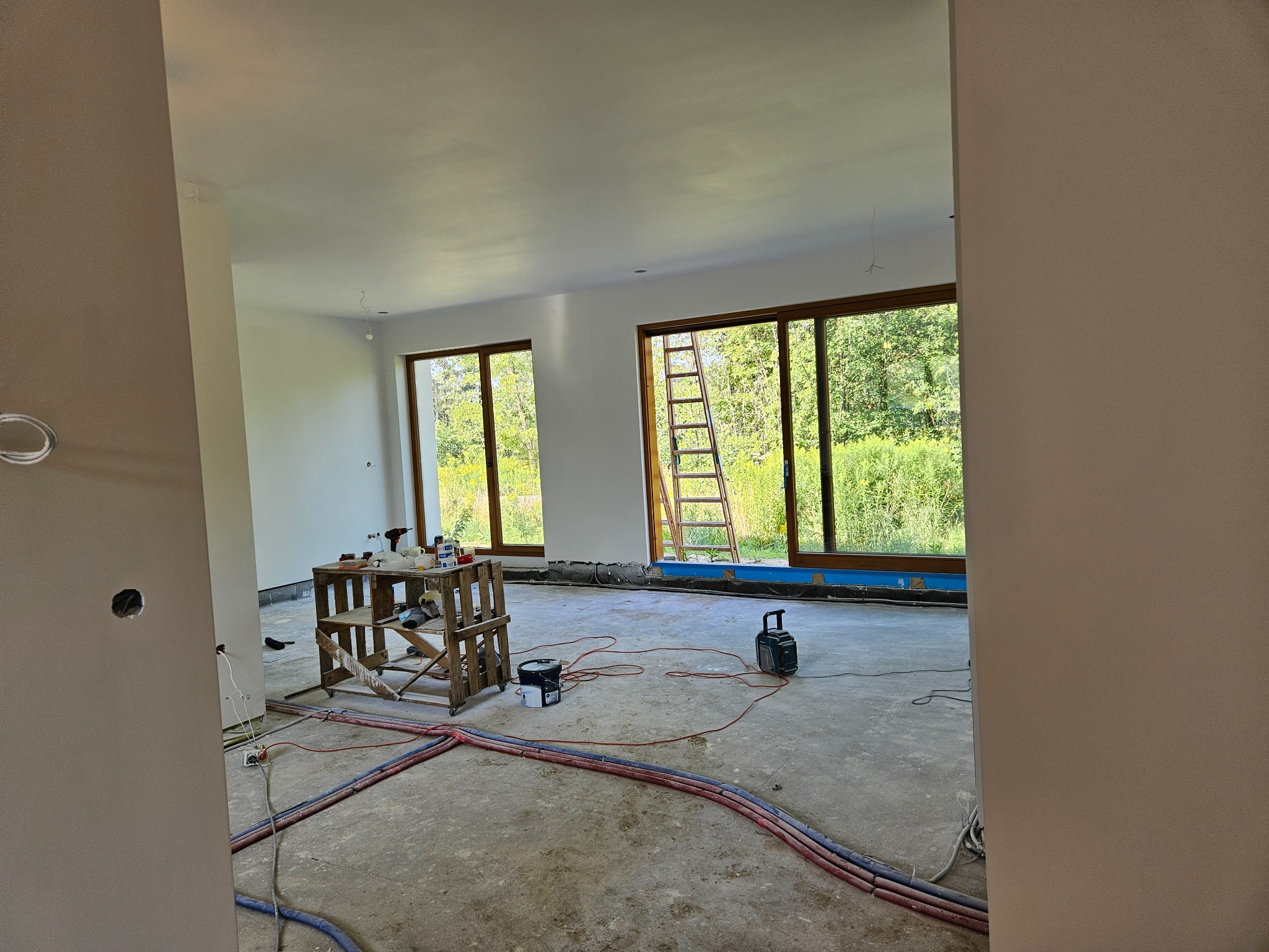 Wnętrze w trakcie prac wykończeniowych: widoczne instalacje podłogowe, drabina oparta o okno, prowizoryczny stół roboczy z narzędziami, ściany przygotowane do malowania.