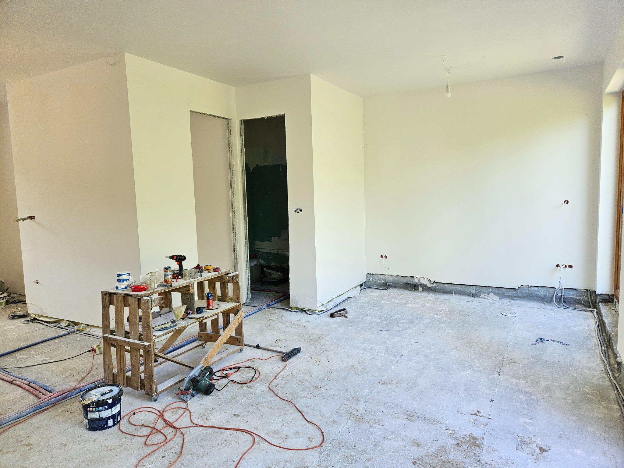 Wnętrze w trakcie remontu: świeżo pomalowane ściany na biało, widoczne wystające kable elektryczne, drewniany podest z narzędziami i farbami, przygotowywane do dalszych prac wykończeniowych.