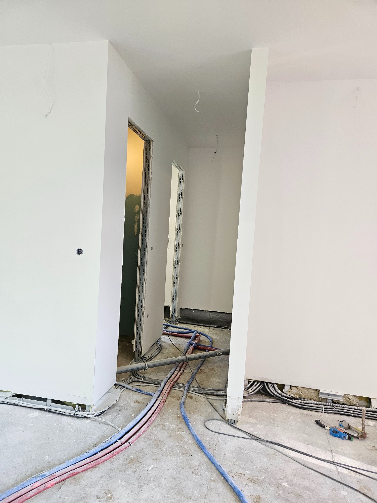 Wnętrze w trakcie prac wykończeniowych: widoczne przewody elektryczne i rury grzewcze na surowym podłożu, ściany pomalowane na biało, przygotowane otwory drzwiowe z widocznymi ościeżnicami.